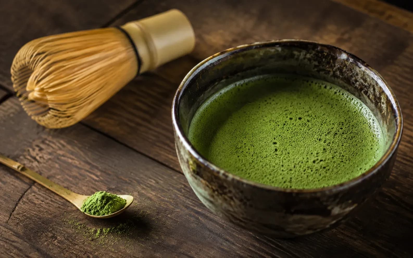 Bowl of freshly whisked matcha beside a bamboo whisk and a scoop of green tea powder.